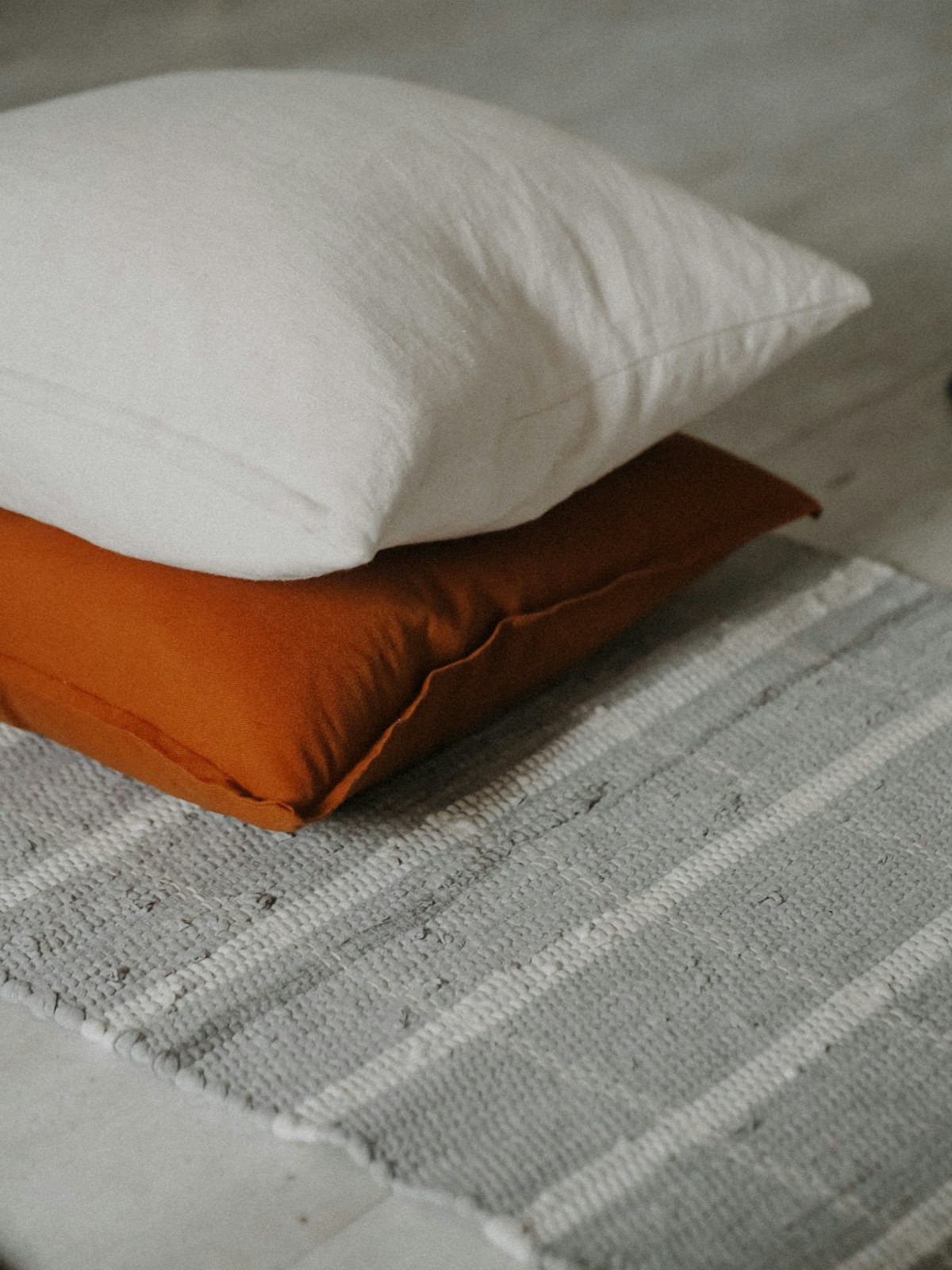 A white linen cushion and a terracotta cushion resting on a woven rug in soft natural light